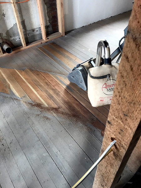 Using a powered floor sander, with a dust collection bag, to sand the floors of the manager's suite. There is a strong contrast betweet the old weathered surface, and the beautiful old growth fir underneath.