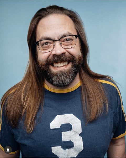 Gabe Rutledge, stand-up comedian. He faces the camera, wears glasses, has long brown hair that hangs below his shoulders, and wears a blue sports jersey with the number 3 in white