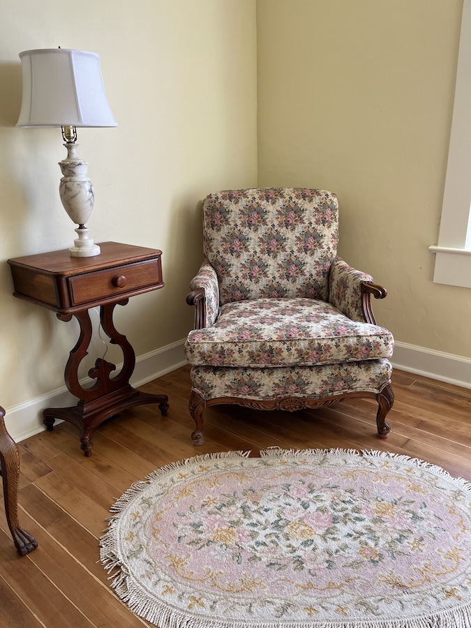 Sitting room of the manager's suite, furnished in period style with a floral upholstered arm chair, a fringed oval rug in a floral pattern, and a vintage ceramic lamp on a vintage phone table.